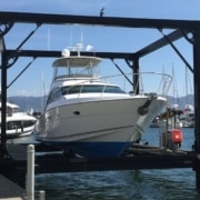Front-facing view of a white and blue motor yacht suspended above the water within a large, black steel boat lift structure. The boat lift is attached to a wooden dock in a sunny marina with many other yachts.