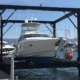 Front-facing view of a white and blue motor yacht suspended above the water within a large, black steel boat lift structure. The boat lift is attached to a wooden dock in a sunny marina with many other yachts.