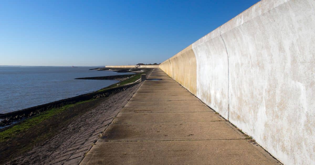 A long, curved concrete sea wall stretching along a coastline under a clear blue sky, designed to redirect wave energy back toward the ocean.
