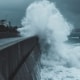 A high, dark gray concrete sea wall structure deflecting a massive, churning white ocean wave during a powerful coastal storm, protecting a road and residential houses on the left.